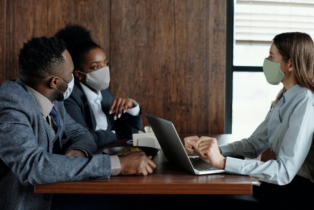 A man and a woman in face masks talking at a table with another woman with a face mask with a laptop.