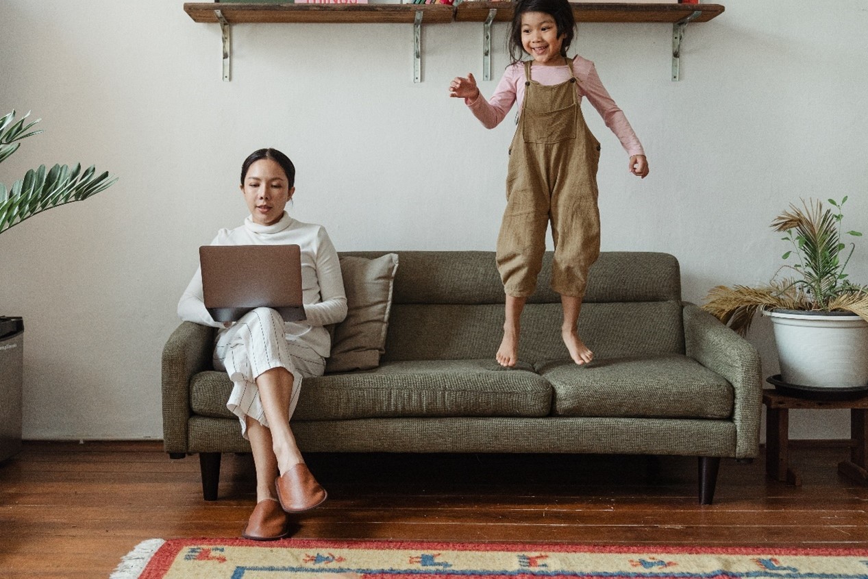 A woman on her laptop working with a child jumping next to her on the couch.