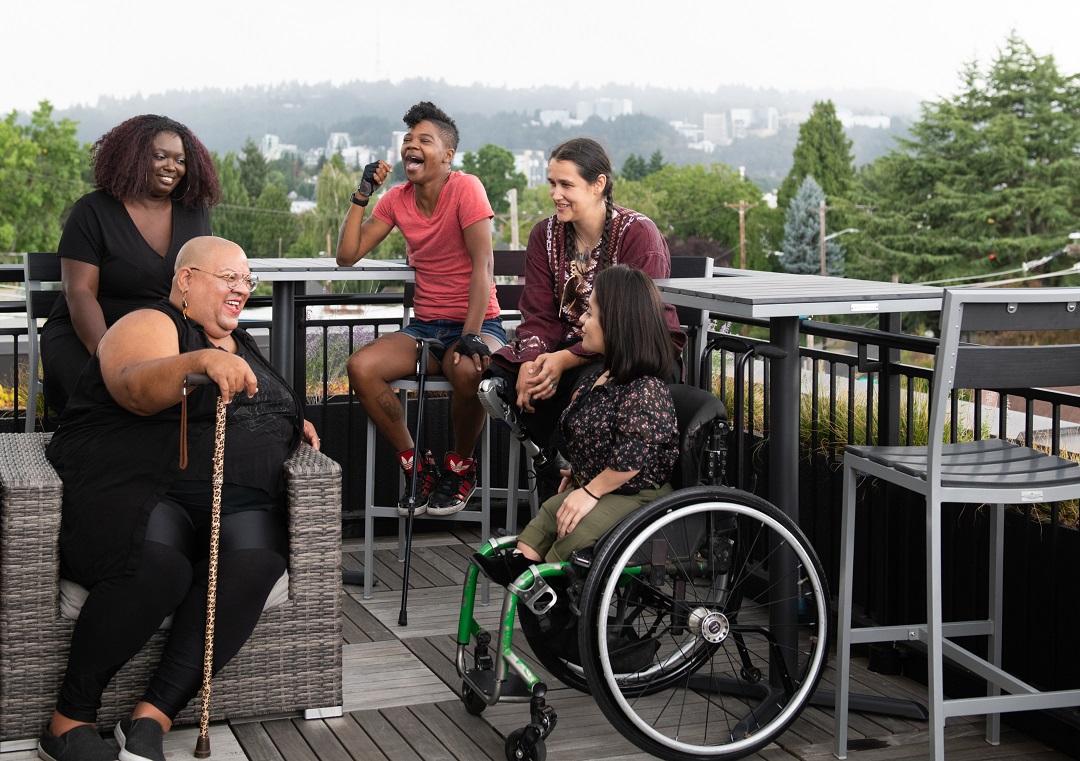 Five disabled people of color with canes, prosthetic legs, and a wheelchair sit on a rooftop deck, laughing and sharing stories. Greenery and city high-rises are visible in the background.