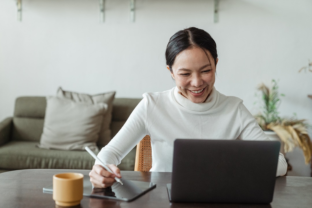 Woman with white turtleneck smiling and looking at computer screen while writing on a pad.