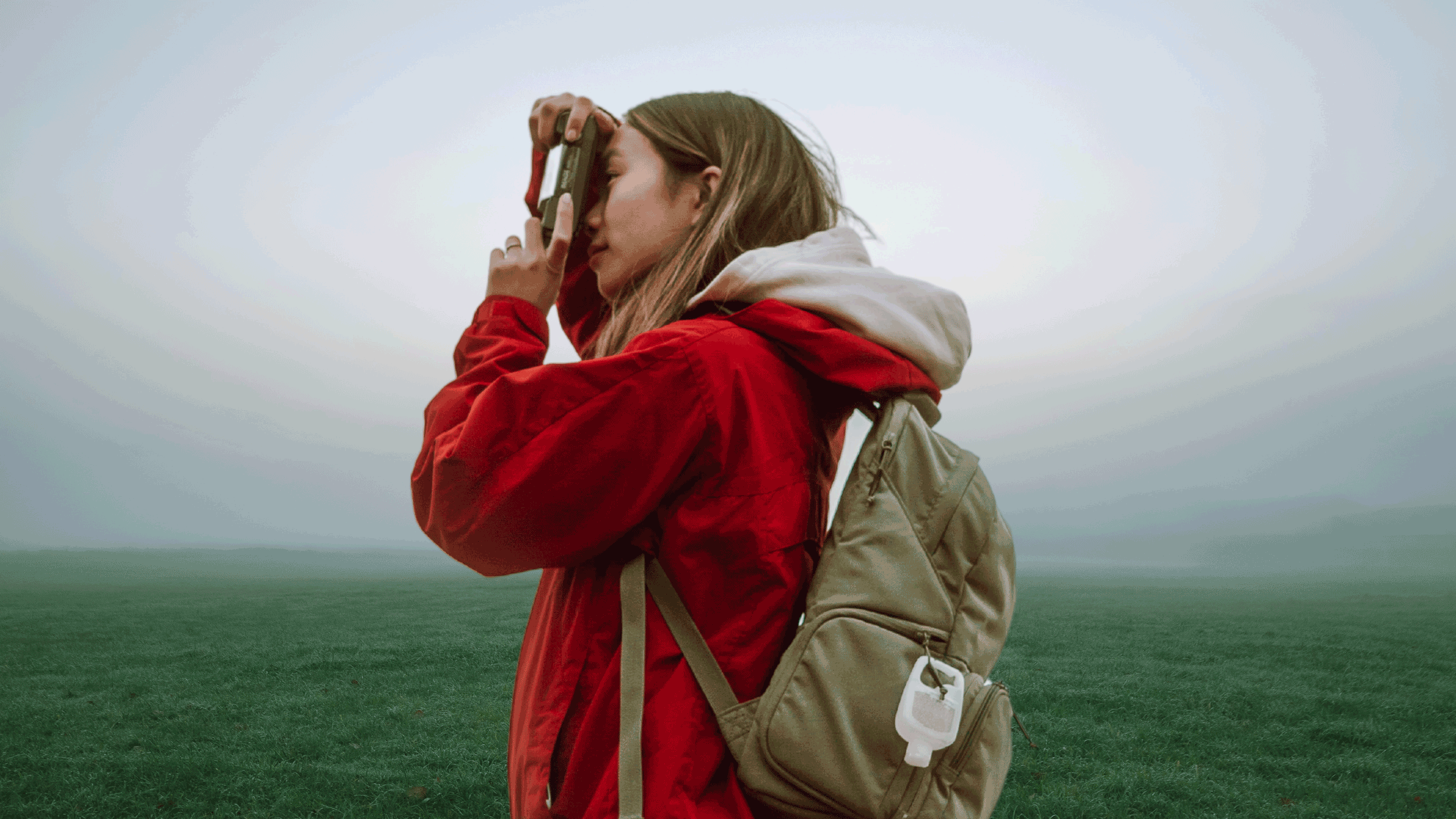Person wearing hoodie and backpack stands outdoors and looks through camera into distance