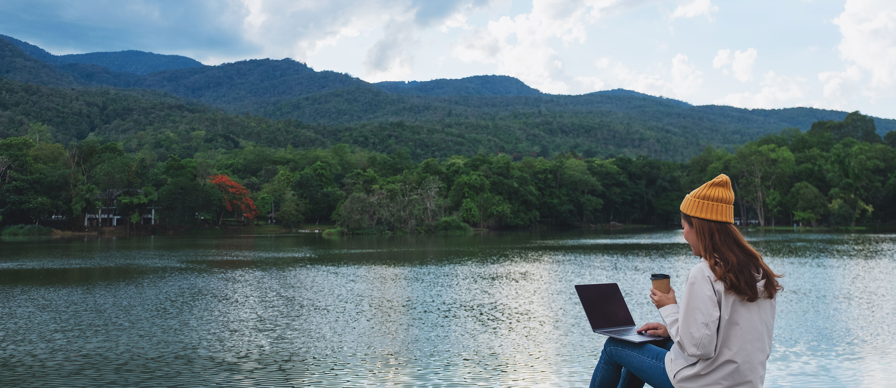 person with laptop sits at edge of mountain lake