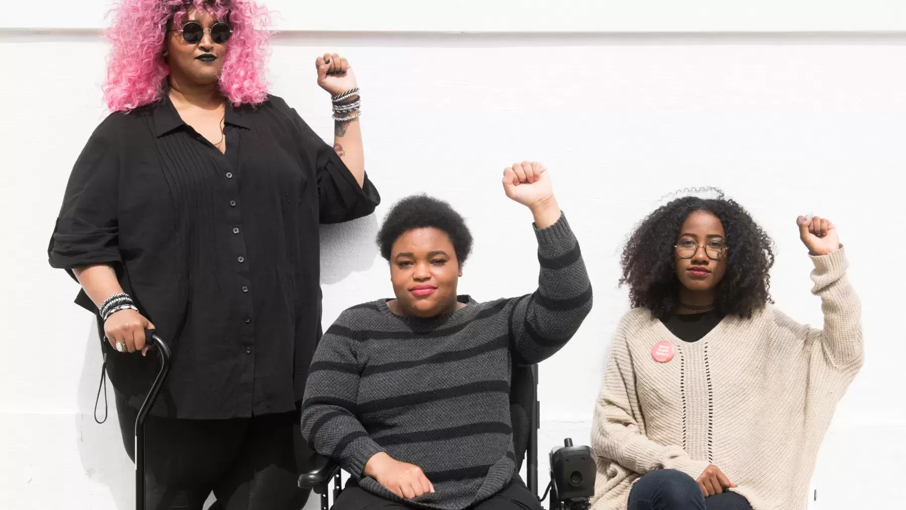 Torso level photo of three Black and disabled folx (a non-binary person holding a cane, a woman in a power wheelchair, and a woman on a folding chair) raising their fists on the sidewalk in front of a white wall.