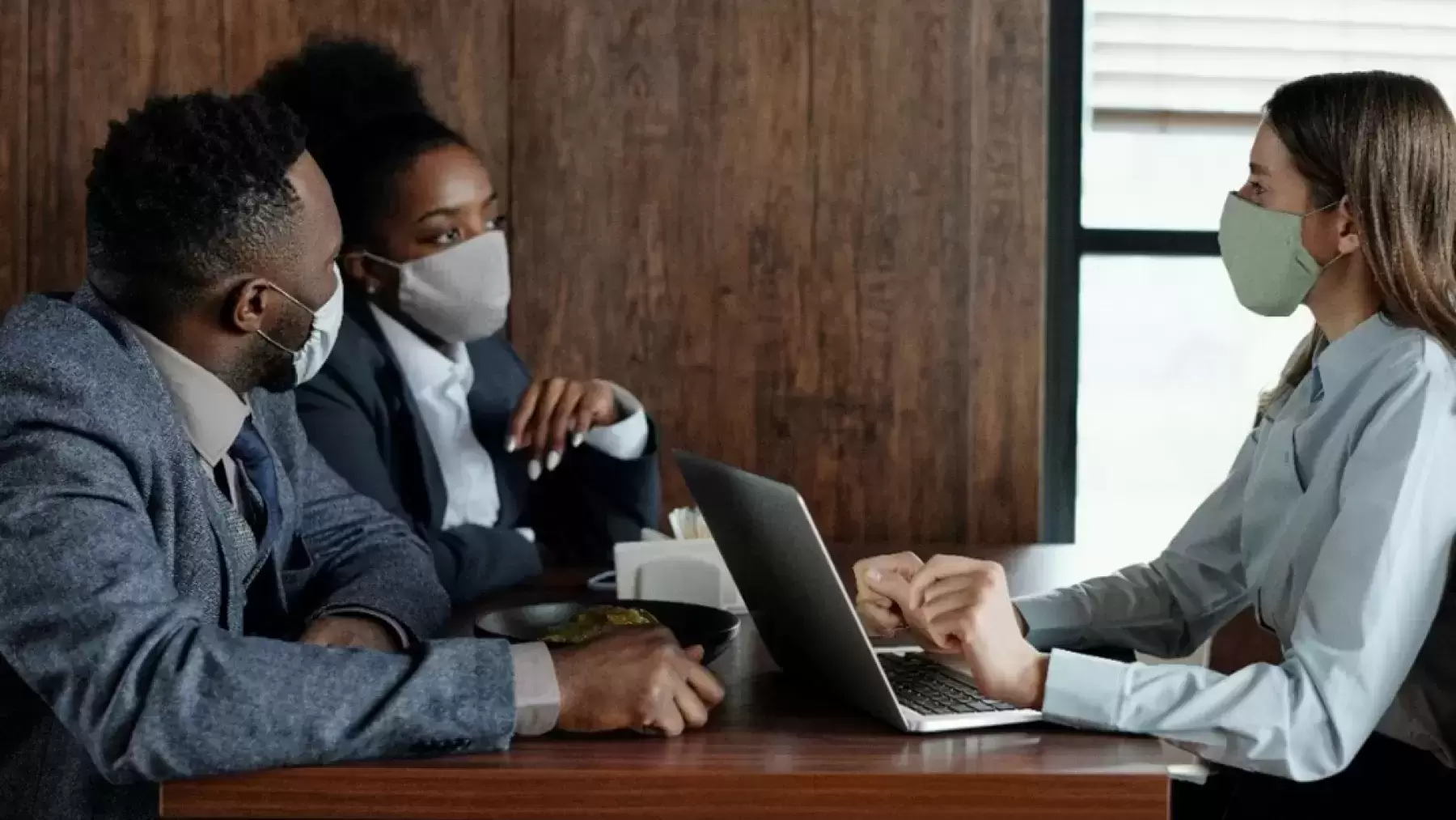 A man and a woman in face masks talking at a table with another woman with a face mask with a laptop.
