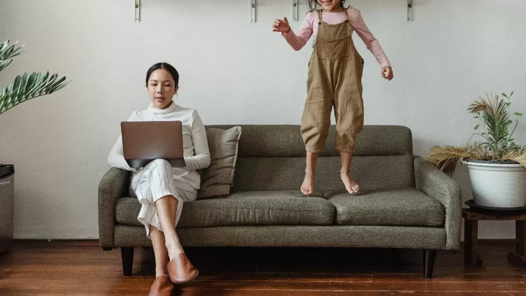 A woman on her laptop working with a child jumping next to her on the couch.