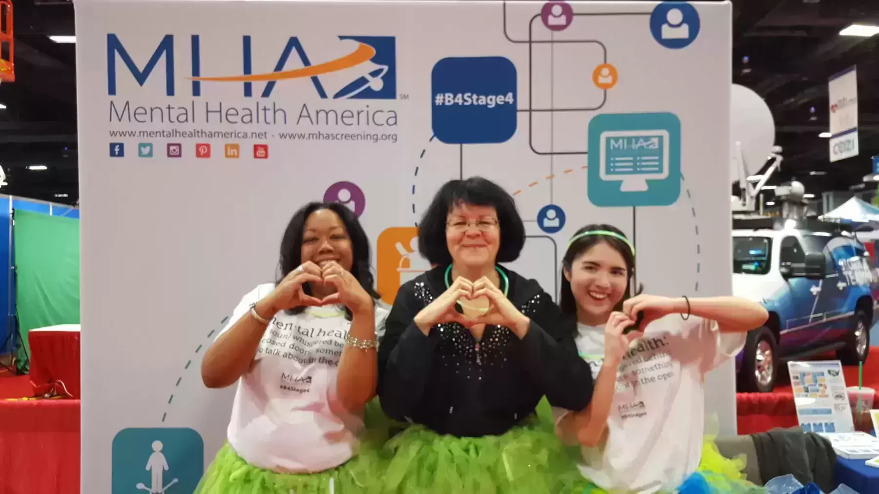 Three women in green tutus posing with their hands in the shapes of hearts in front of an MHA banner