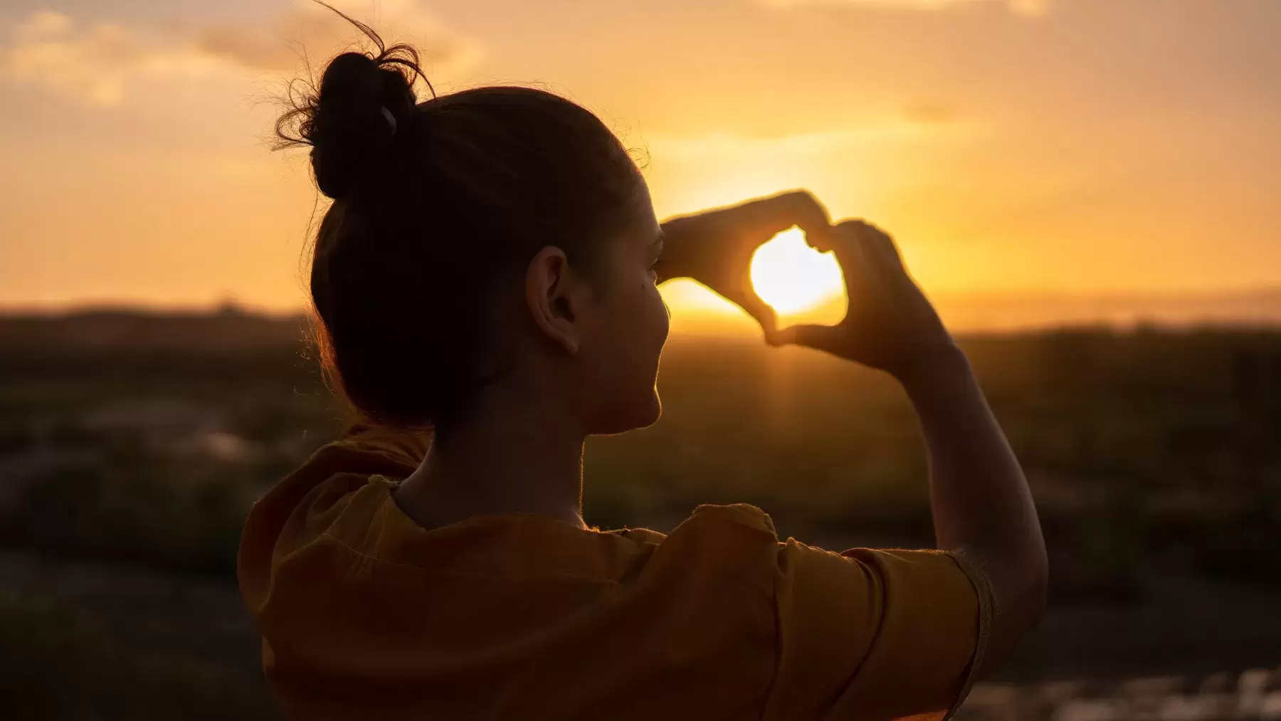 person holds up hands in shape of heart in front of sunset
