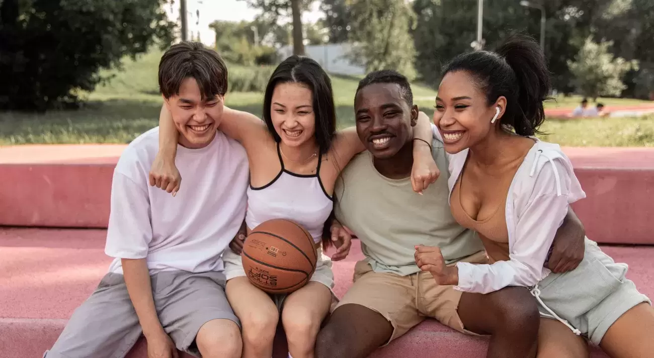 four people in workout clothes sit with arms around each other on a concrete bench smiling, one has a basketball in their lap