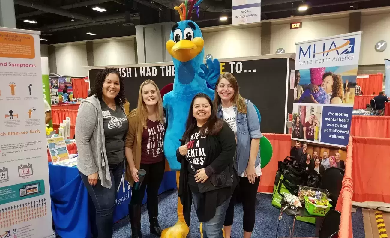 MHA staff and volunteers stand with person in large blue bird costume surrounded by MHA posters and paraphernalia 