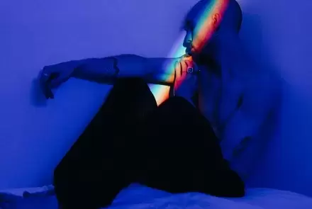 blue shaded image of person on bed leaning in corner of room with rainbow prism reflecting on their face