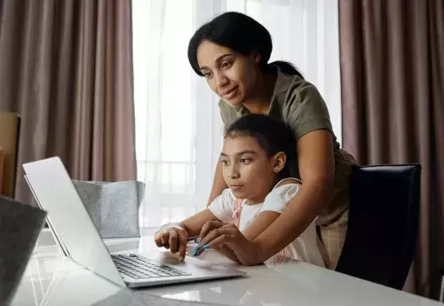 woman leans over child who is sitting while both look at computer screen together at a table