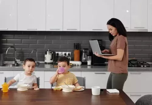 woman holds a computer and works on it while walking behind two children who eat at a table