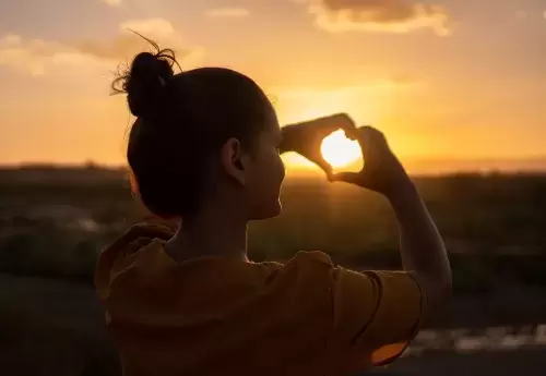 person holds up hands in shape of heart in front of sunset