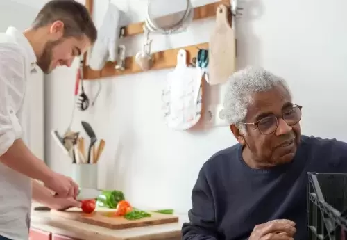 younger person standing chops vegetables while older person sits