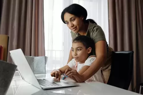 woman leans over child who is sitting while both look at computer screen together at a table