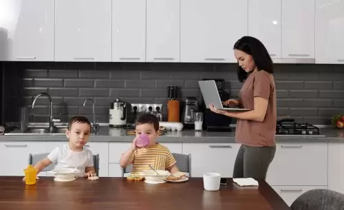 woman holds a computer and works on it while walking behind two children who eat at a table