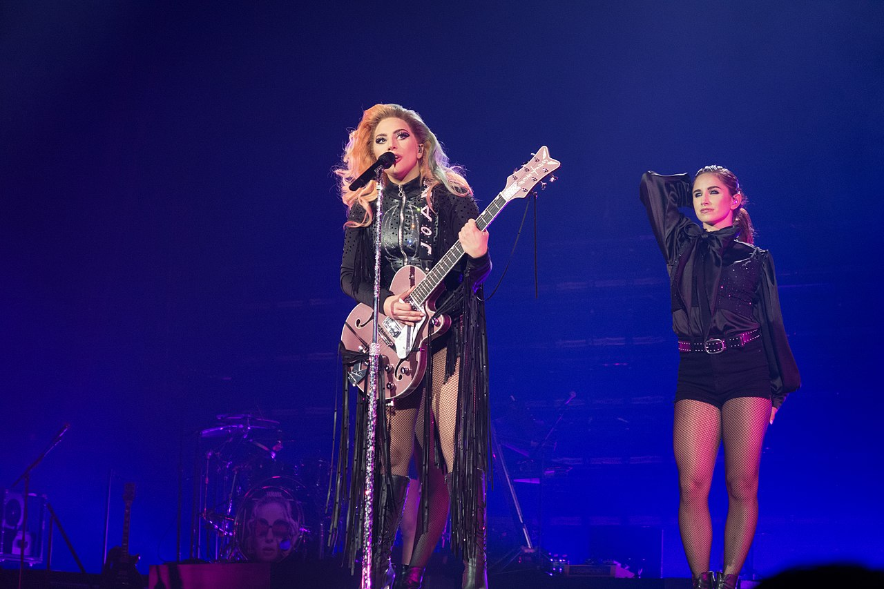 Lada Gaga stands on stage at microphone holding guitar with backup dancer behind her